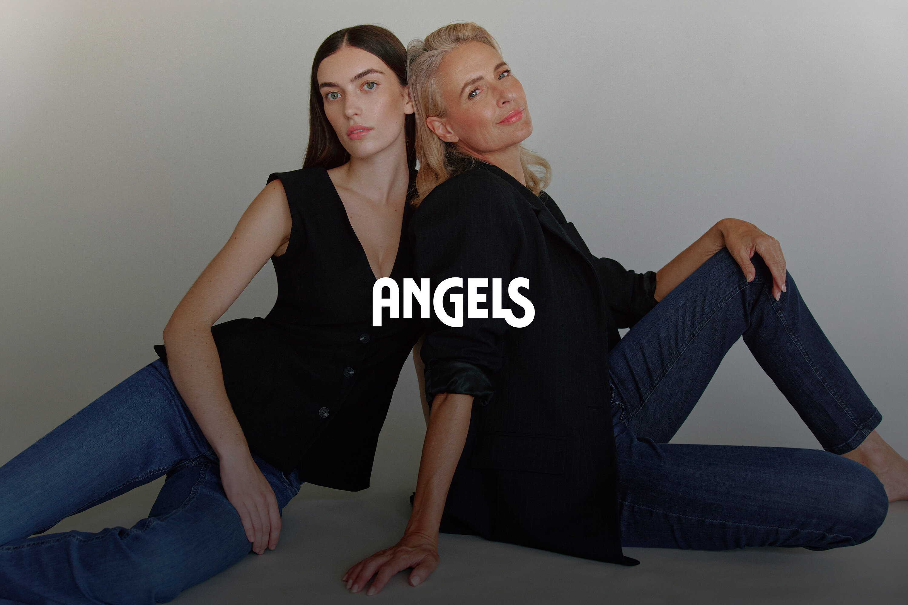 Dwie kobiety siedzą tyłem do siebie. Młodsza kobieta w czarnej kamizelce i dżinsach, starsza kobieta w czarnej marynarce i dżinsach.