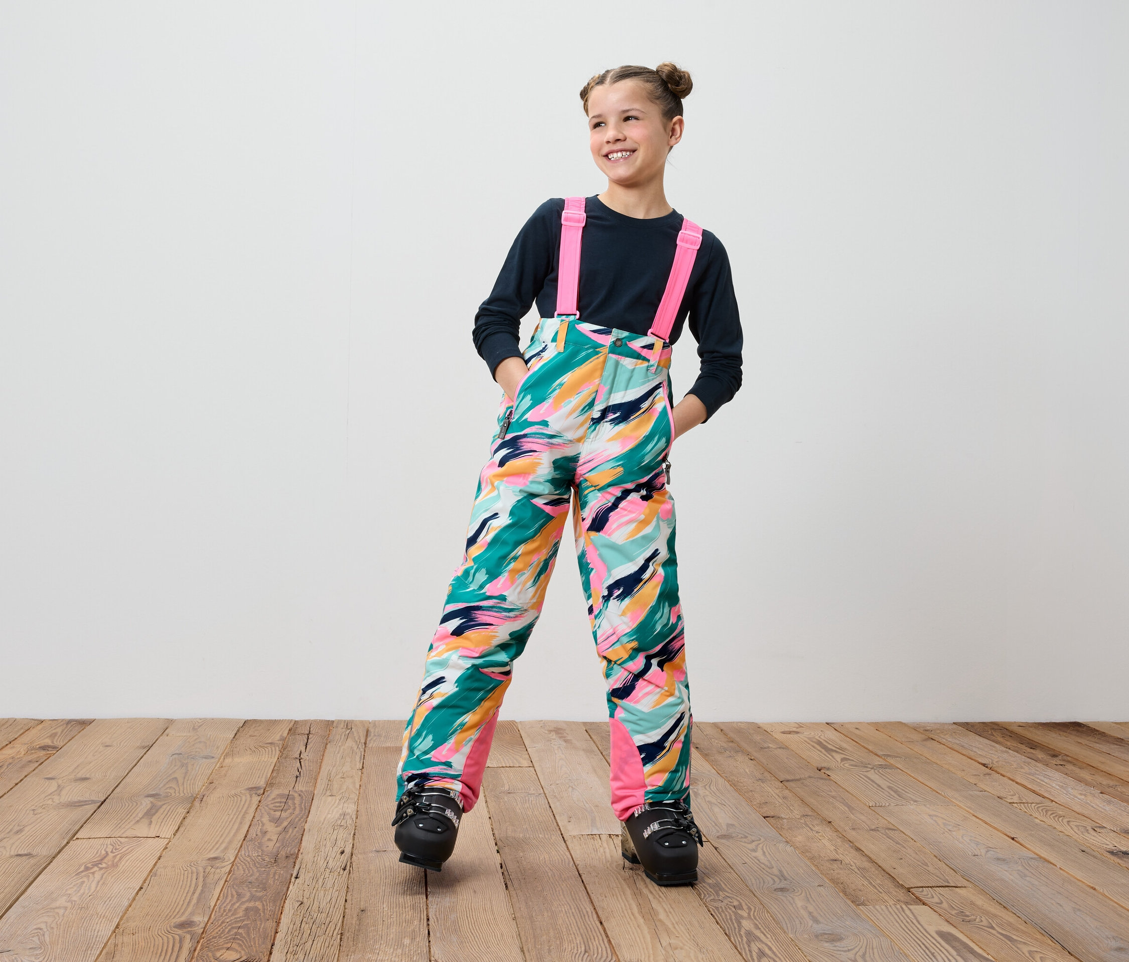 Uśmiechnięta nastolatka w kolorowych spodniach narciarskich na szelkach i czarnych butach narciarskich stoi na drewnianej podłodze.