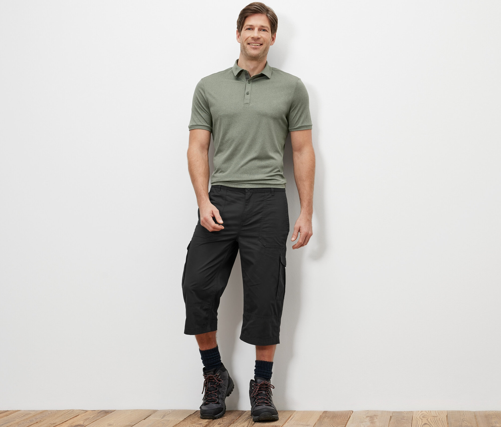 Mężczyzna stoi i ma na sobie zieloną koszulkę polo i antracytowe bermudy bojówki o długości 3/4.