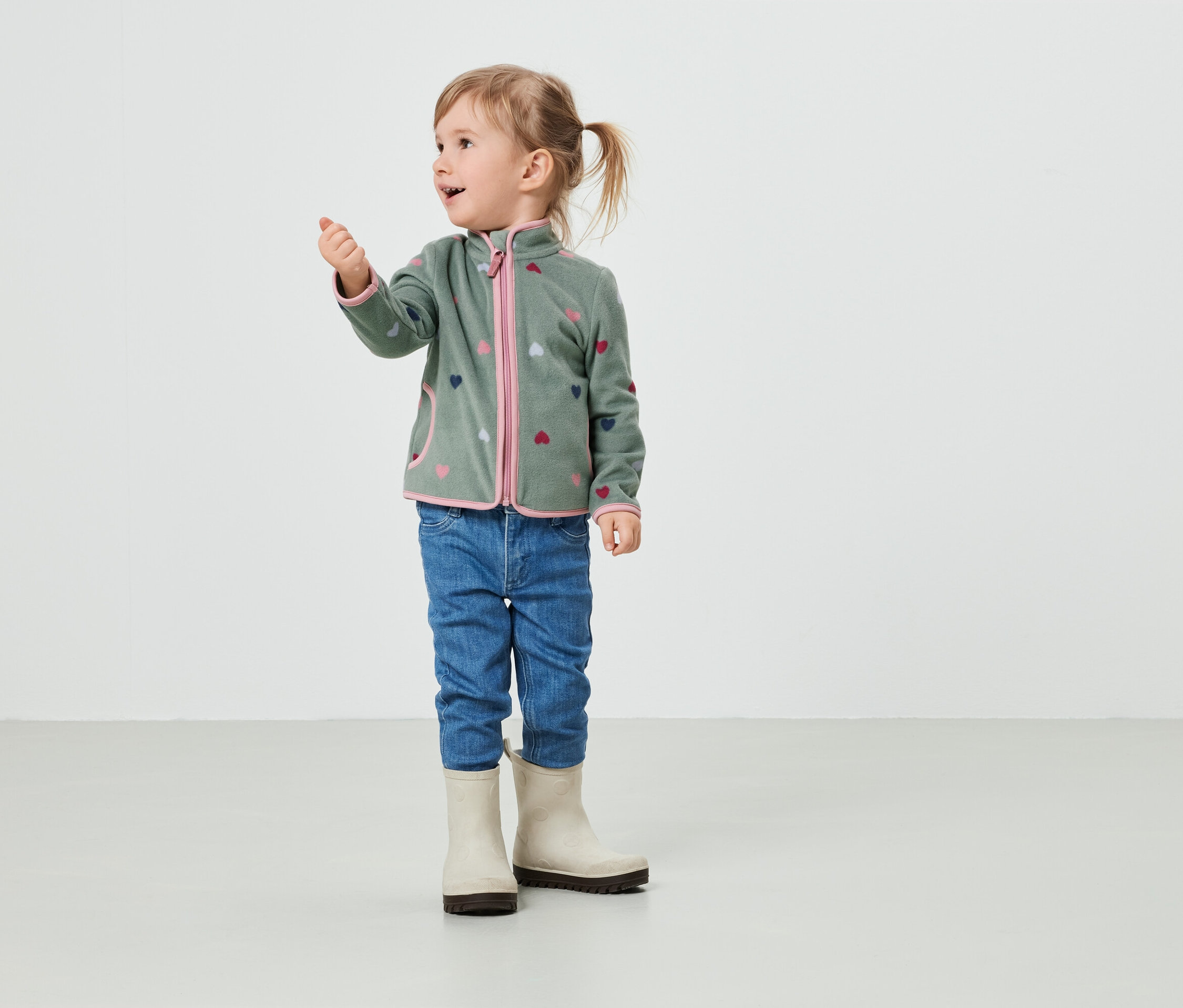 Dziewczynka w oliwkowozielonej kurtce polarowej w serduszka, dżinsach i kaloszach stoi w pokoju.