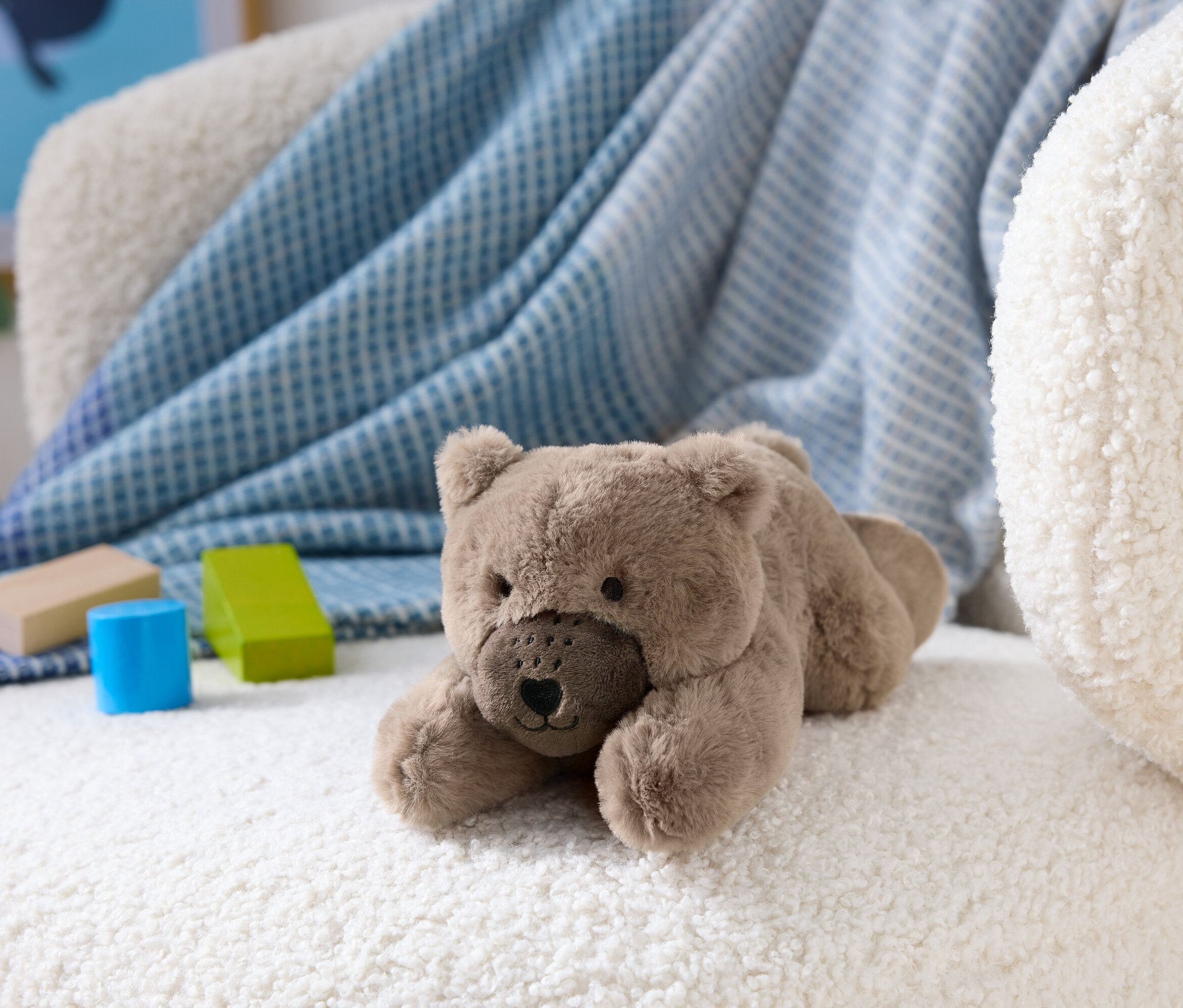 Brązowy pluszowy miś leży na białym, puszystym krześle obok drewnianych klocków.