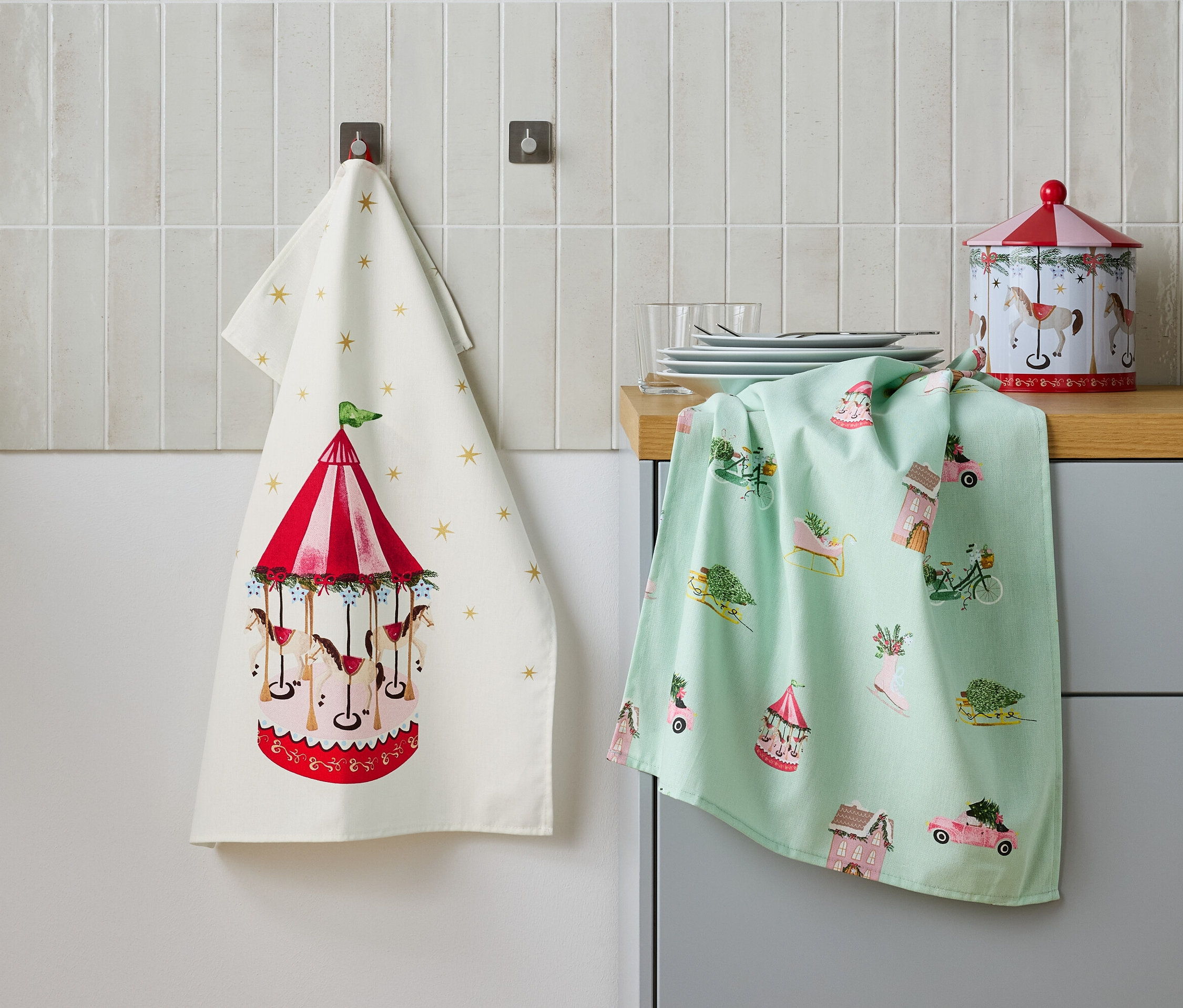 Dwie ściereczki wiszą obok siebie na ścianie, jedna z motywem karuzeli, a druga z motywami świątecznymi, oraz metalowa puszka na stole.