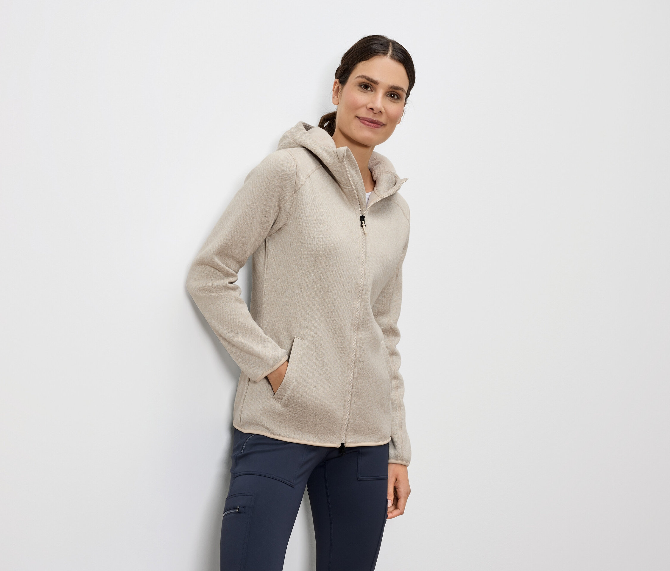 Kobieta stoi z ręką w kieszeni beżowej bluzy z kapturem na zamek i niebieskich spodniach.