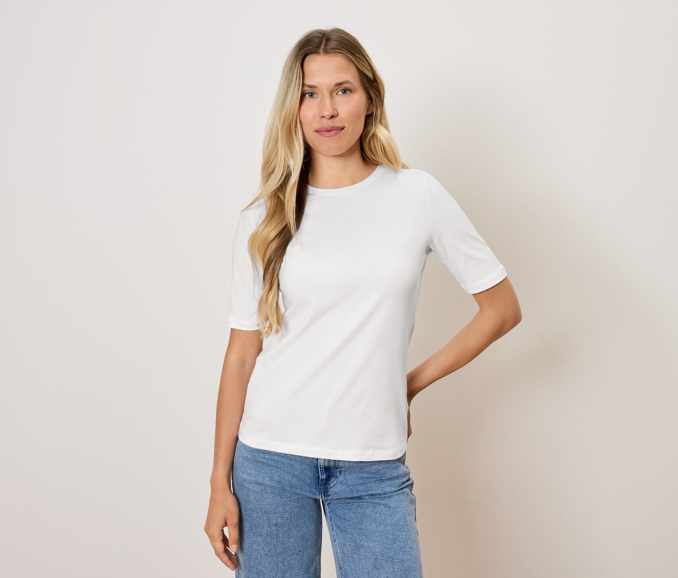 Kobieta o blond włosach pozuje w białej koszulce i niebieskich dżinsach.