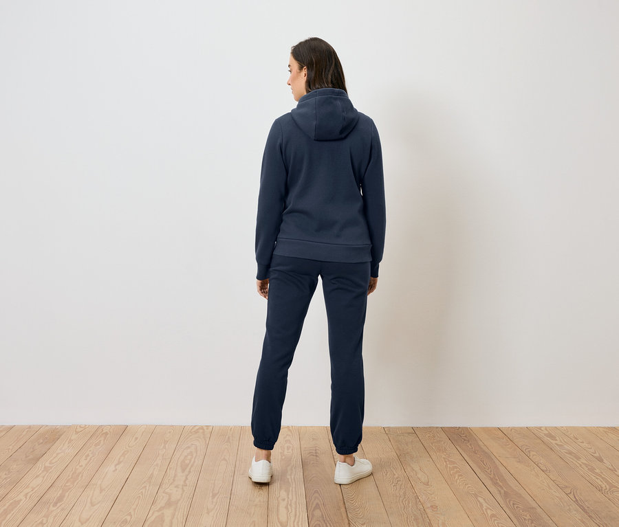 Kobieta od tyłu w niebieskiej bluzie z kapturem i spodniach stoi na drewnianej podłodze przed białą ścianą.