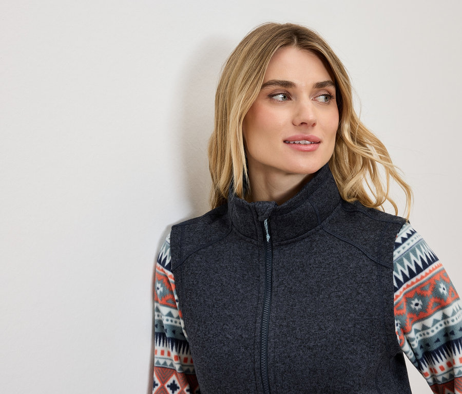 Blondynka w ciemnoszarej kamizelce z ozdobnym wzorem na rękawach patrzy w lewo.