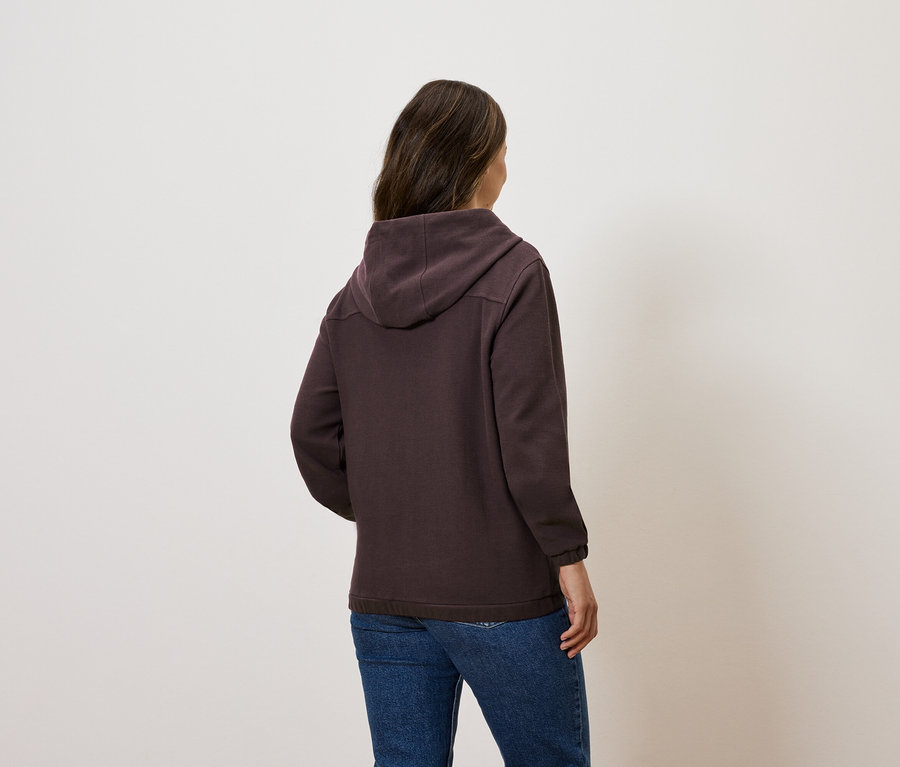 Kobieta stoi tyłem do kamery, ubrana w brązową bluzę z kapturem i dżinsy.