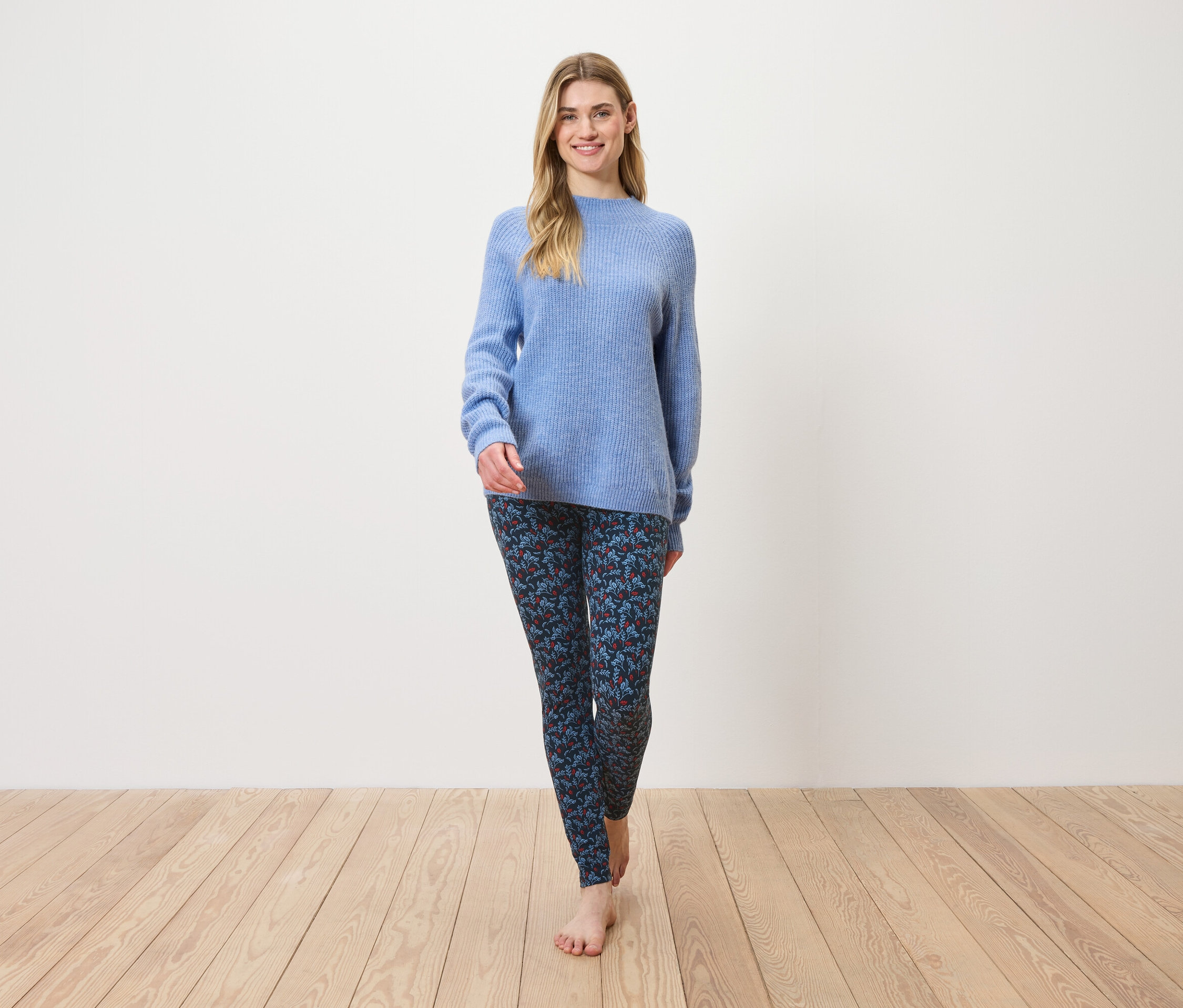 Kobieta idzie boso po drewnianej podłodze, ubrana w niebieski sweter i wzorzyste legginsy.