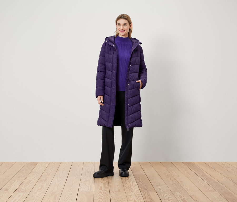 Kobieta stoi na drewnianej podłodze, ubrana w fioletowy płaszcz, fioletowy sweter i czarne spodnie.