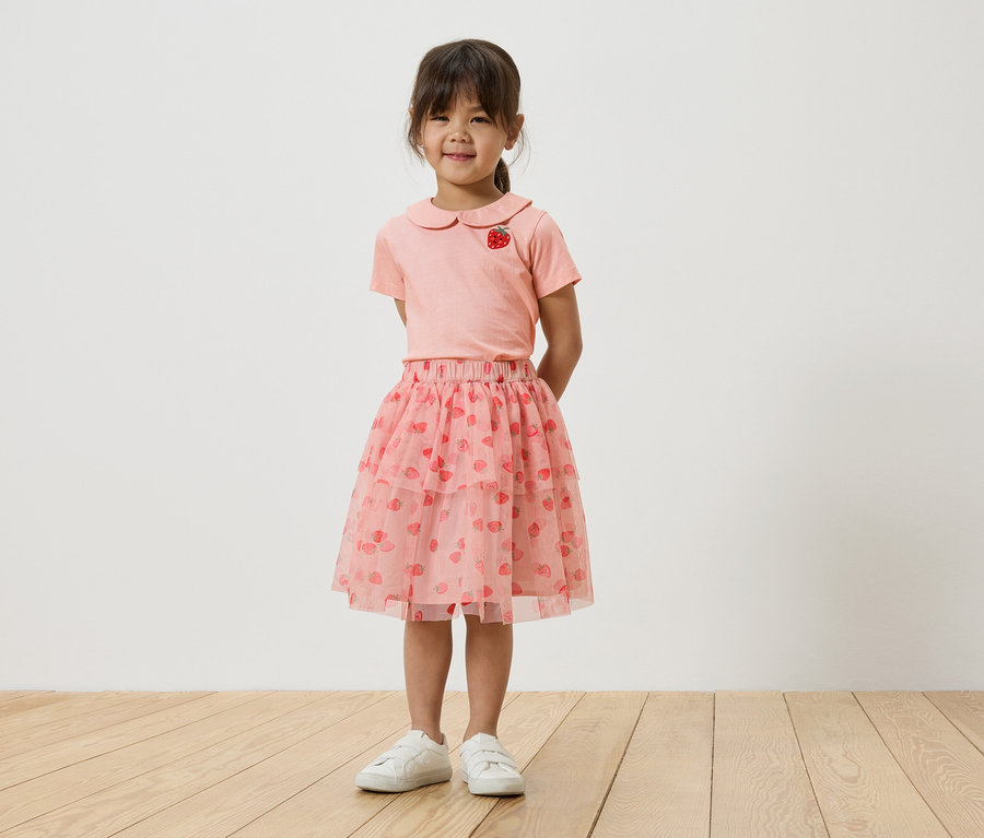 Dziewczynka stoi na tle białej ściany, ubrana w różowy T-shirt z motywem truskawki, różową tiulową spódniczkę w truskawki i białe tenisówki.