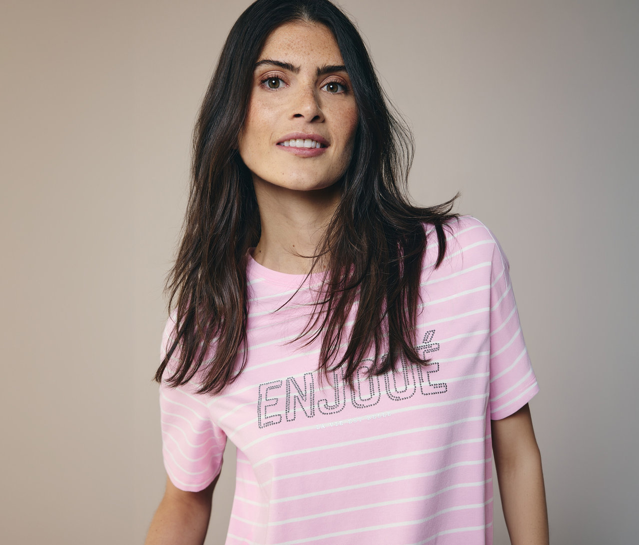 Kobieta z długimi, ciemnymi włosami ma na sobie koszulkę CECIL w różowo-białe paski z napisem 'ENJOUE'.
