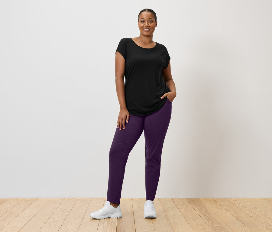 Kobieta w czarnym T-shircie, fioletowych spodniach i białych butach sportowych stoi na tle białej ściany.