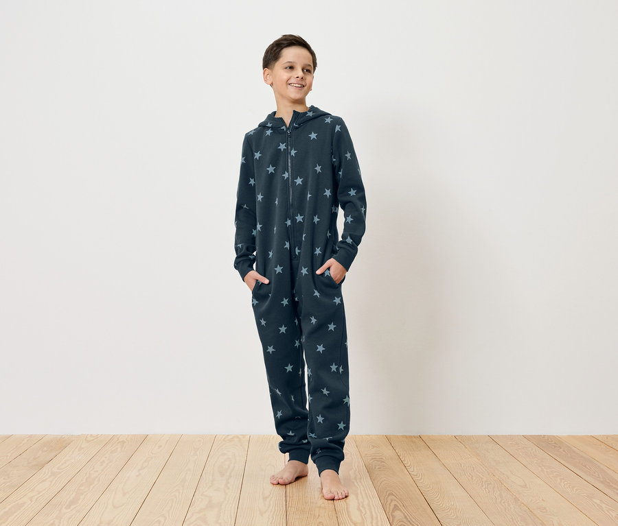 Młody mężczyzna stoi boso na drewnianej podłodze, ubrany w granatowy kombinezon w gwiazdy z kapturem i zamkiem błyskawicznym, z rękami w kieszeniach.