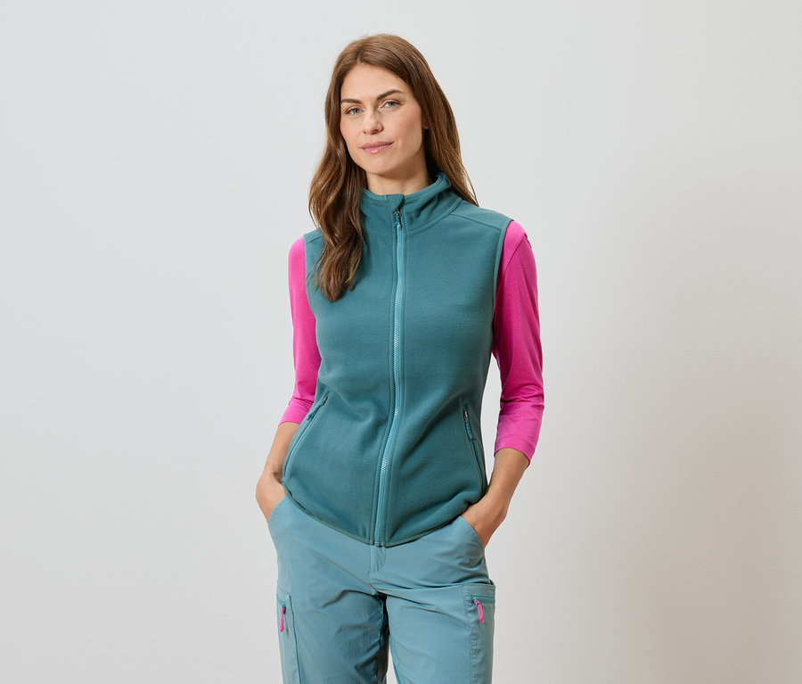 Kobieta w petrol barwy kamizelce, dopasowanych spodniach i różowej bluzce z długim rękawem.