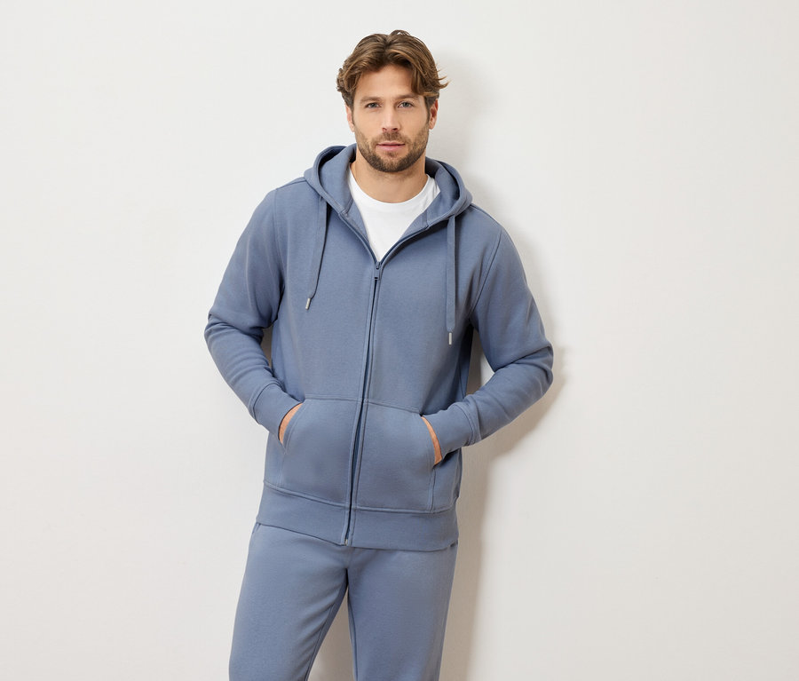 Mężczyzna o jasnobrązowych włosach w niebieskim dresie z kapturem i suwakiem oraz białym t-shircie, stoi z obiema rękami w kieszeniach.