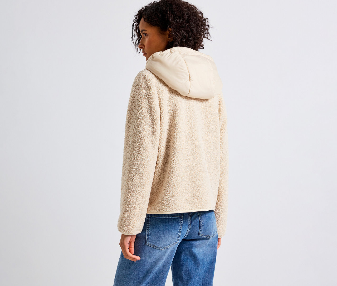 Kobieta ma na sobie bezową bluzę pluszową z odpinanym kapturem marki Street One i niebieskie dżinsy.