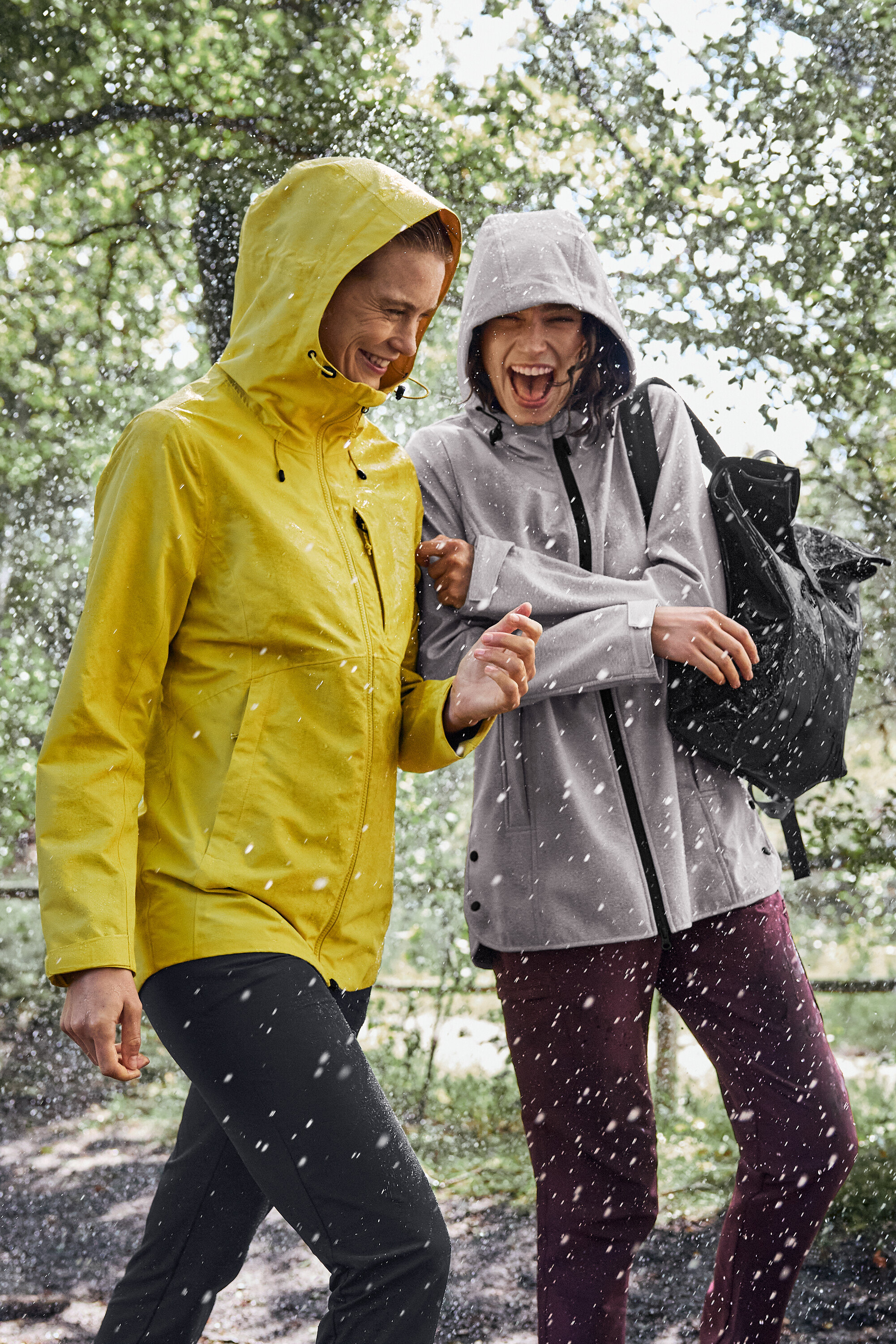 Dwie kobiety idą w deszczu, jedna ma na sobie żółtą kurtkę przeciwdeszczową, druga szarą z czarnym plecakiem.