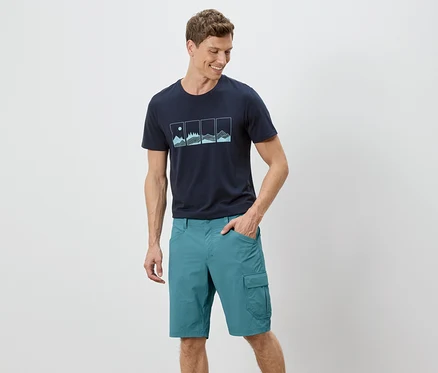 Mężczyzna stoi z ręką w kieszeni na białym tle. Ma na sobie granatowy T-shirt z grafiką i zielone szorty.