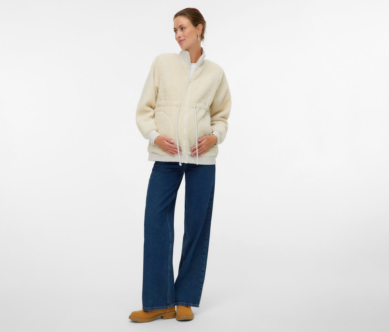 Kobieta w ciąży ma na sobie pluszową bluzę 3 w 1 MAMALICIOUS „MLCARLY”, dżinsy i botki.