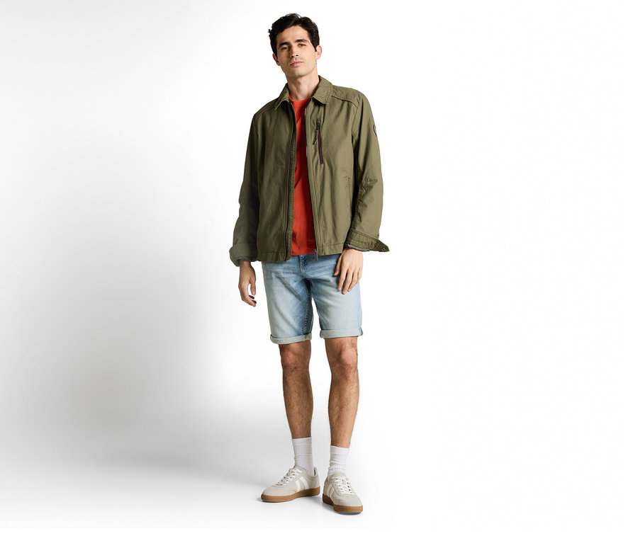 Mężczyzna w oliwkowej kurtce i szortach dżinsowych Tom Tailor TTJOSH REGULAR SLIM, light stone blue denim, do tego T-shirt, skarpety i tenisówki.