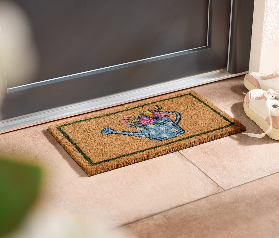 Wycieraczka z motywem konewki leży przed drzwiami, obok stoją białe trampki.