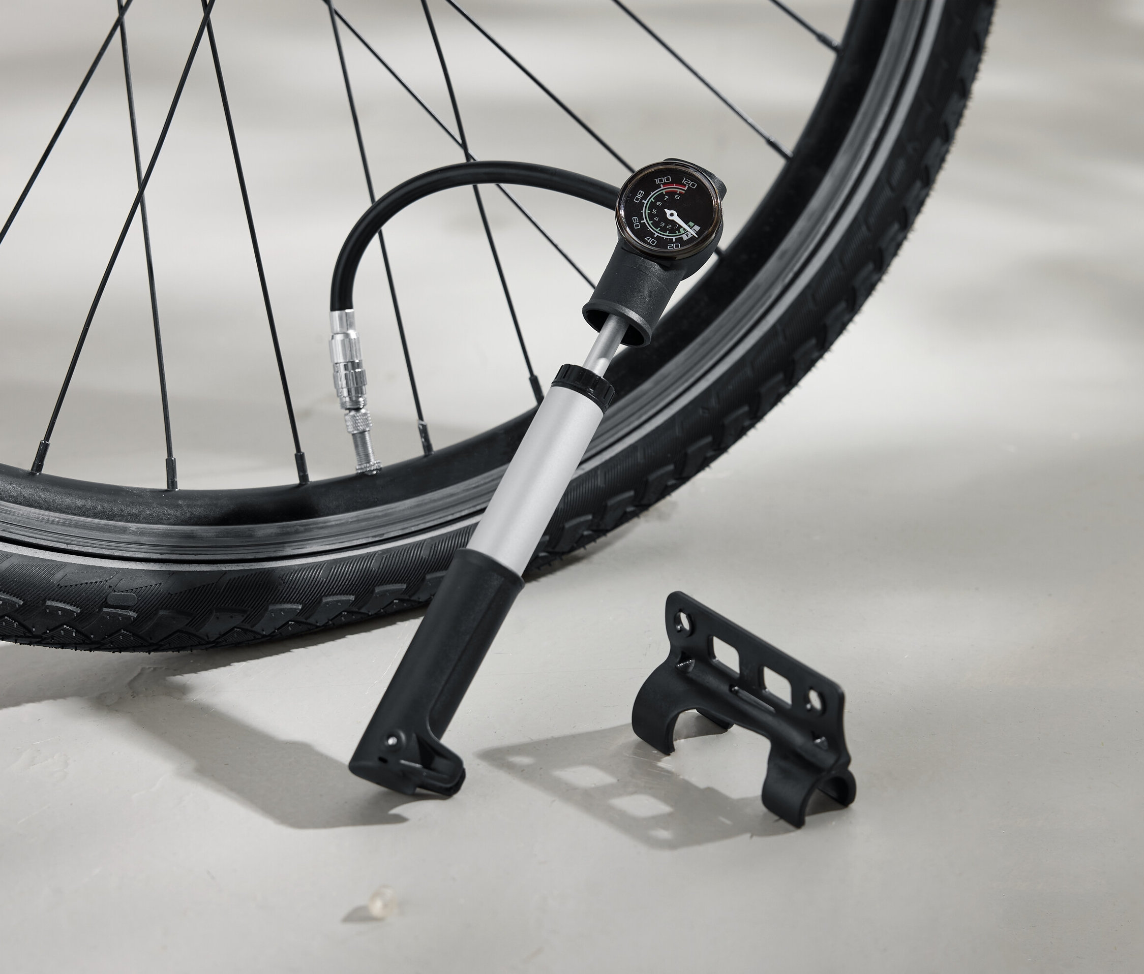 Opona rowerowa pompowana srebrną pompką z czarną rączką i analogowym manometrem. Obok na ziemi leży czarny wspornik.