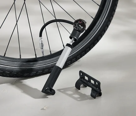 Opona rowerowa pompowana srebrną pompką z czarną rączką i analogowym manometrem. Obok na ziemi leży czarny wspornik.