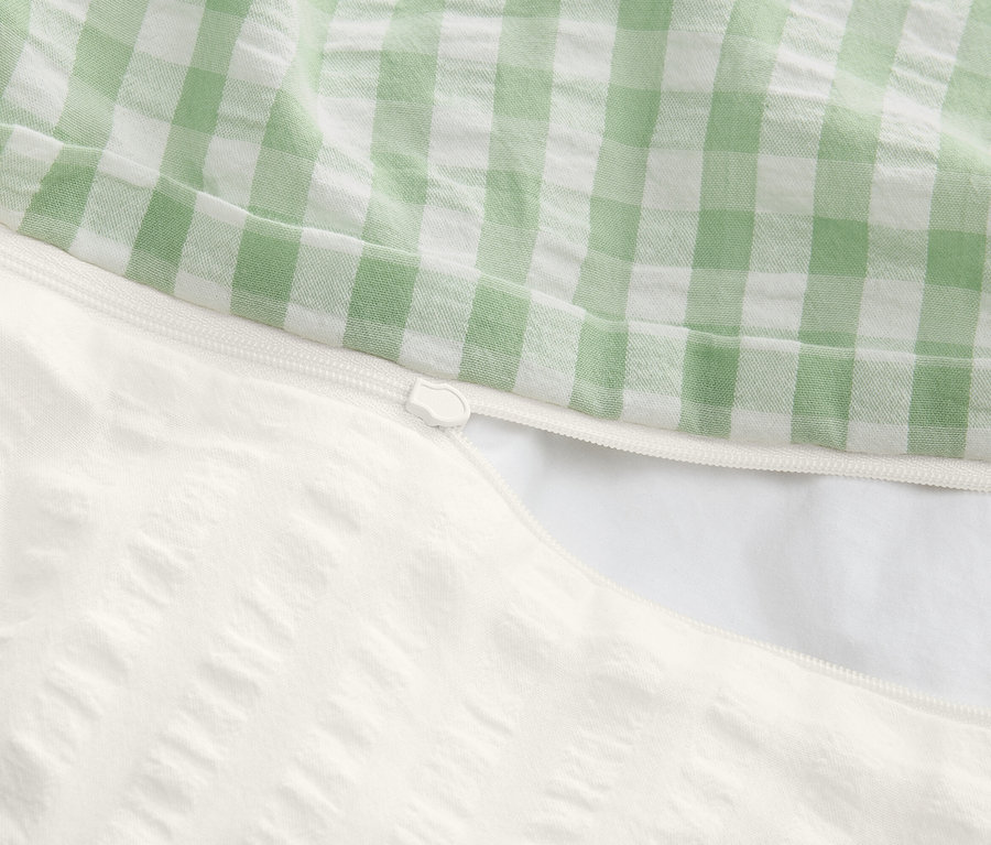 Zbliżenie częściowo otwartej poszwy na kołdrę z zamkiem błyskawicznym, góra w zielono-białą kratę, dół biały z teksturą.