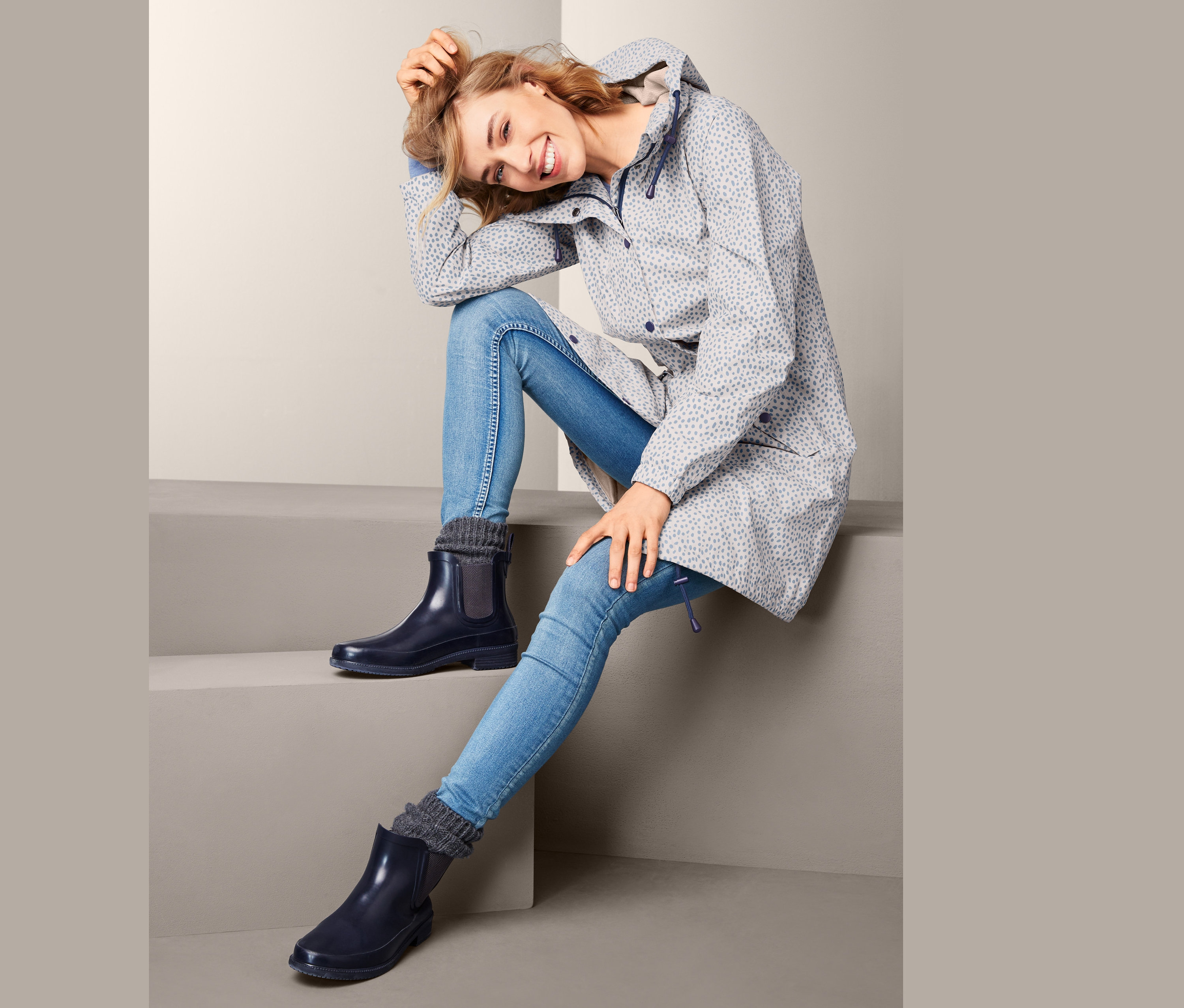 Kobieta siedzi na schodach w płaszczu przeciwdeszczowym i niebieskich kaloszach.