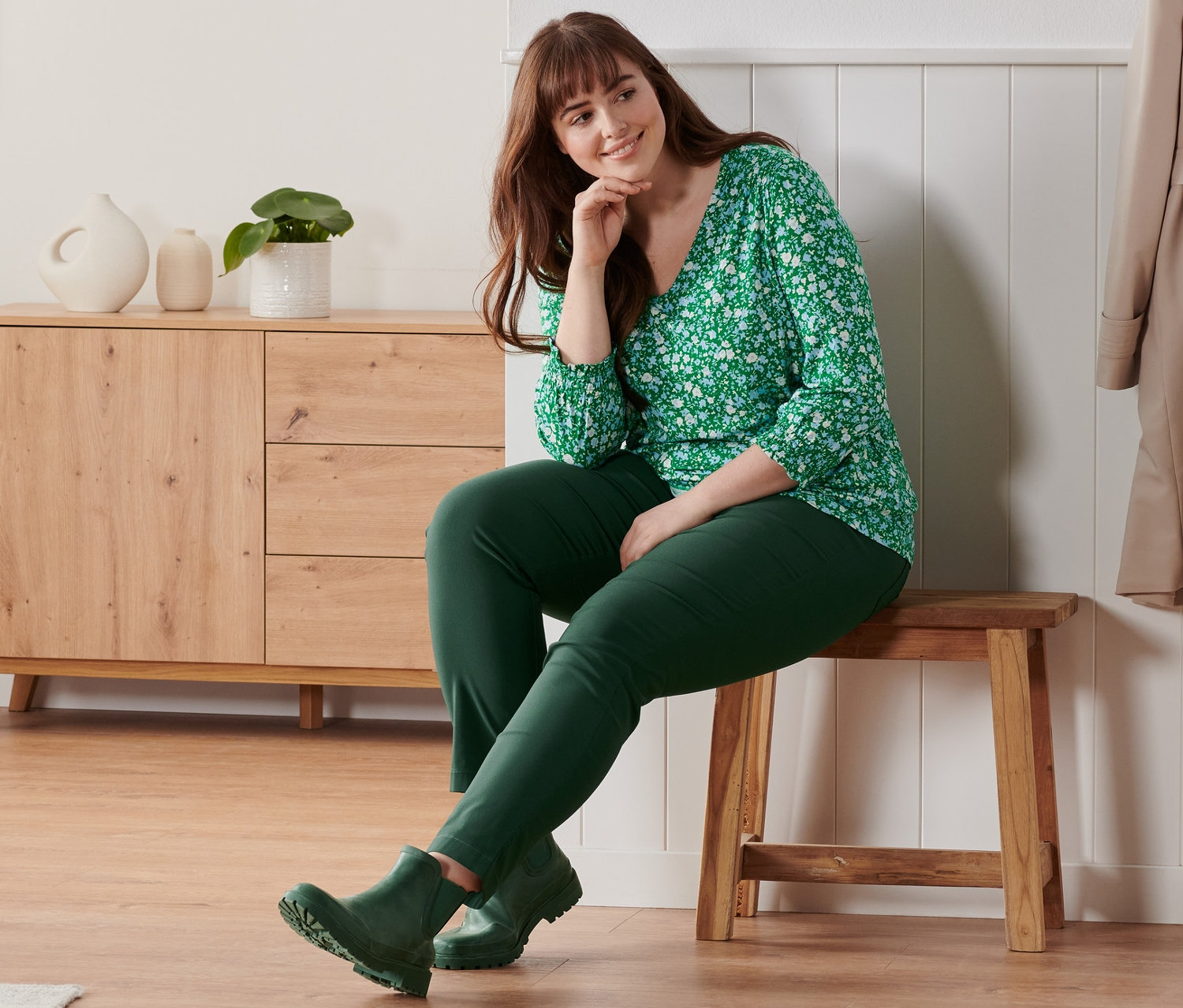 Kobieta siedzi na ławce i ma na sobie zieloną bluzkę z rękawami o długości 3/4 z kwiatowym nadrukiem na całej powierzchni, spodnie z bengaliny i botki-kalosze.