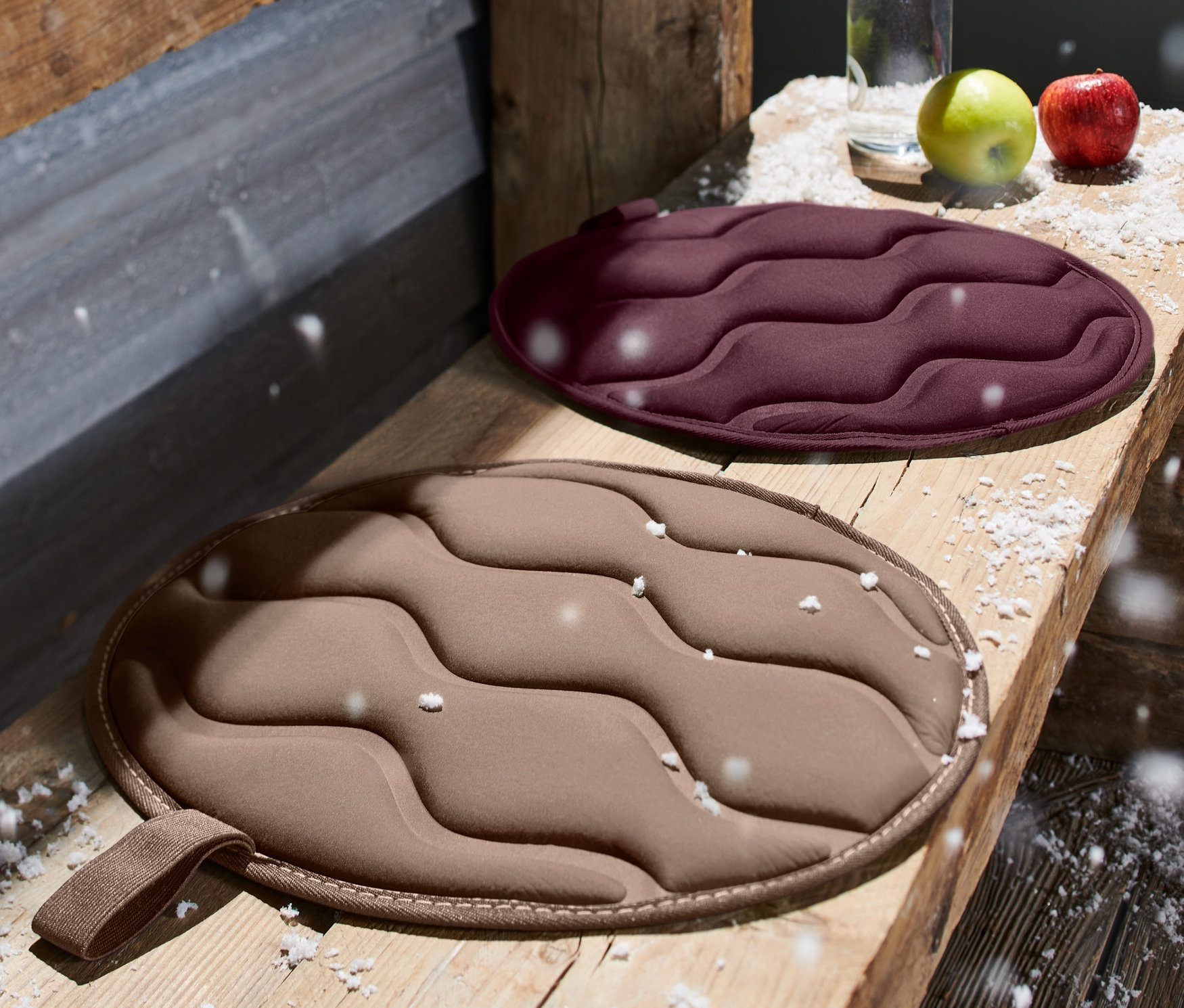 Dwie okrągłe, składane poduszki termiczne do siedzenia leżą na drewnianej ławce. Jedna jest bordowa, a druga beżowa.