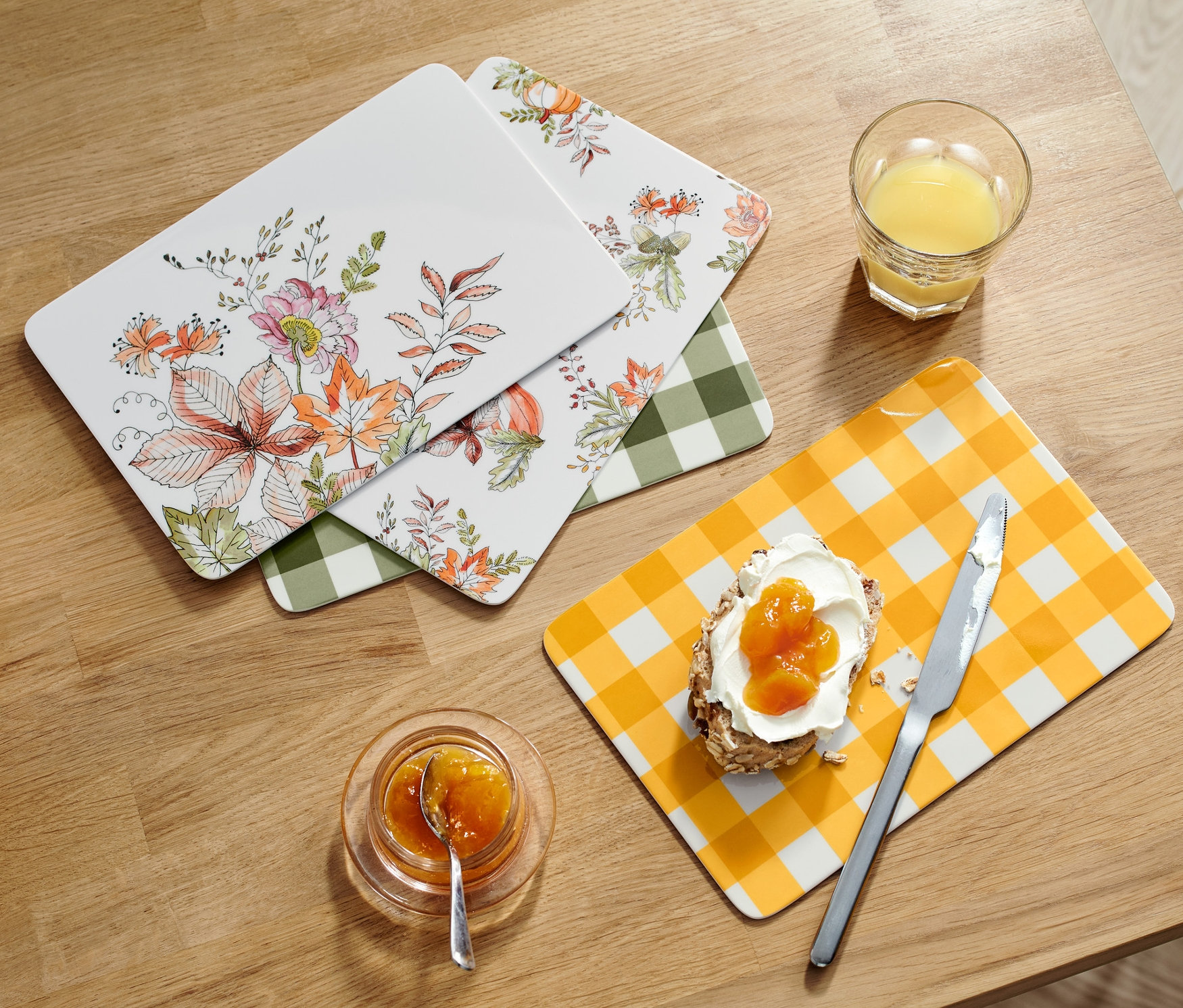 Cztery deski śniadaniowe leżą na drewnianym stole obok szklanki soku, chleba z dżemem, dżemu i koszyka z chlebem.