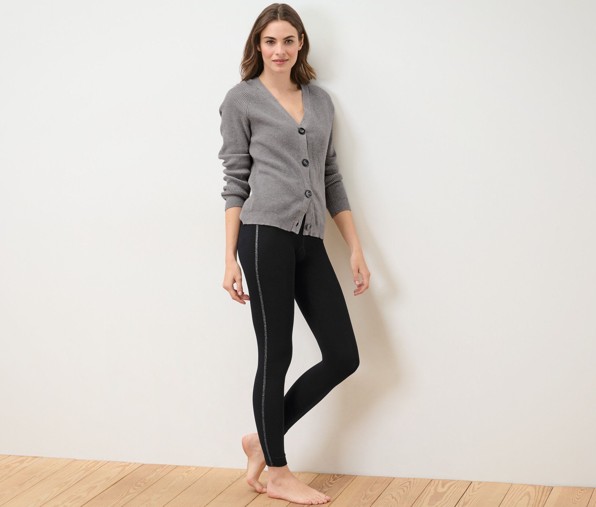 Kobieta pozuje w legginsach termoaktywnych z błyszczącą taśmą i szarym kardiganie.