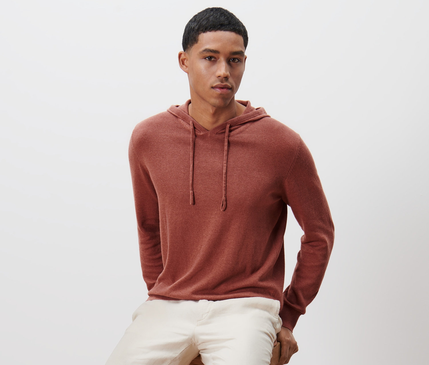 Mężczyzna siedzi na drewnianym stołku i ma na sobie dzianinową bluzę z kapturem.