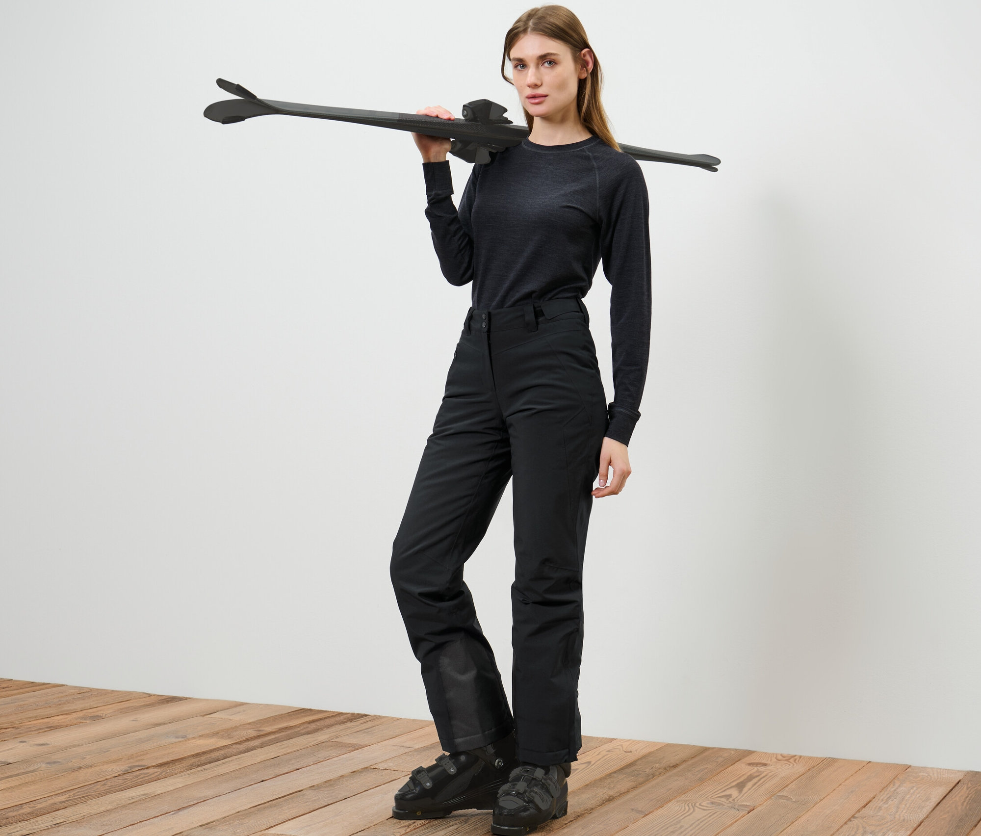 Kobieta pozuje w spodniach narciarskich, trzymając narty na ramieniu.