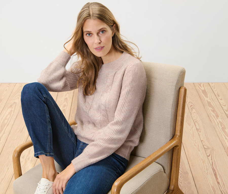 Kobieta siedzi w fotelu i ma na sobie sweter o grubym splocie z wzorem warkoczowym.
