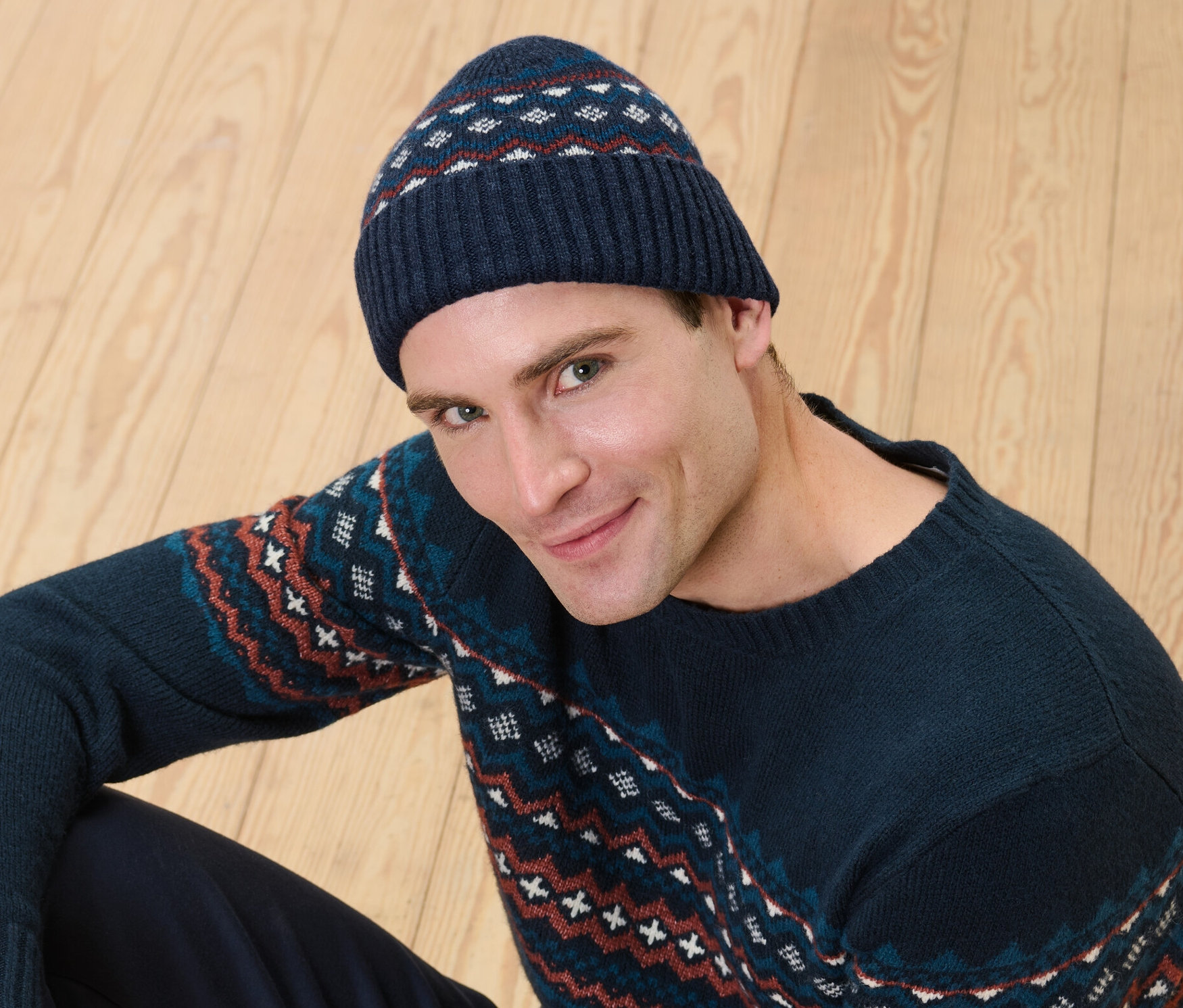 Mężczyzna siedzi na drewnianej podłodze, ma na sobie czapkę z wełną z certyfikatem RWS i sweter we wzory.