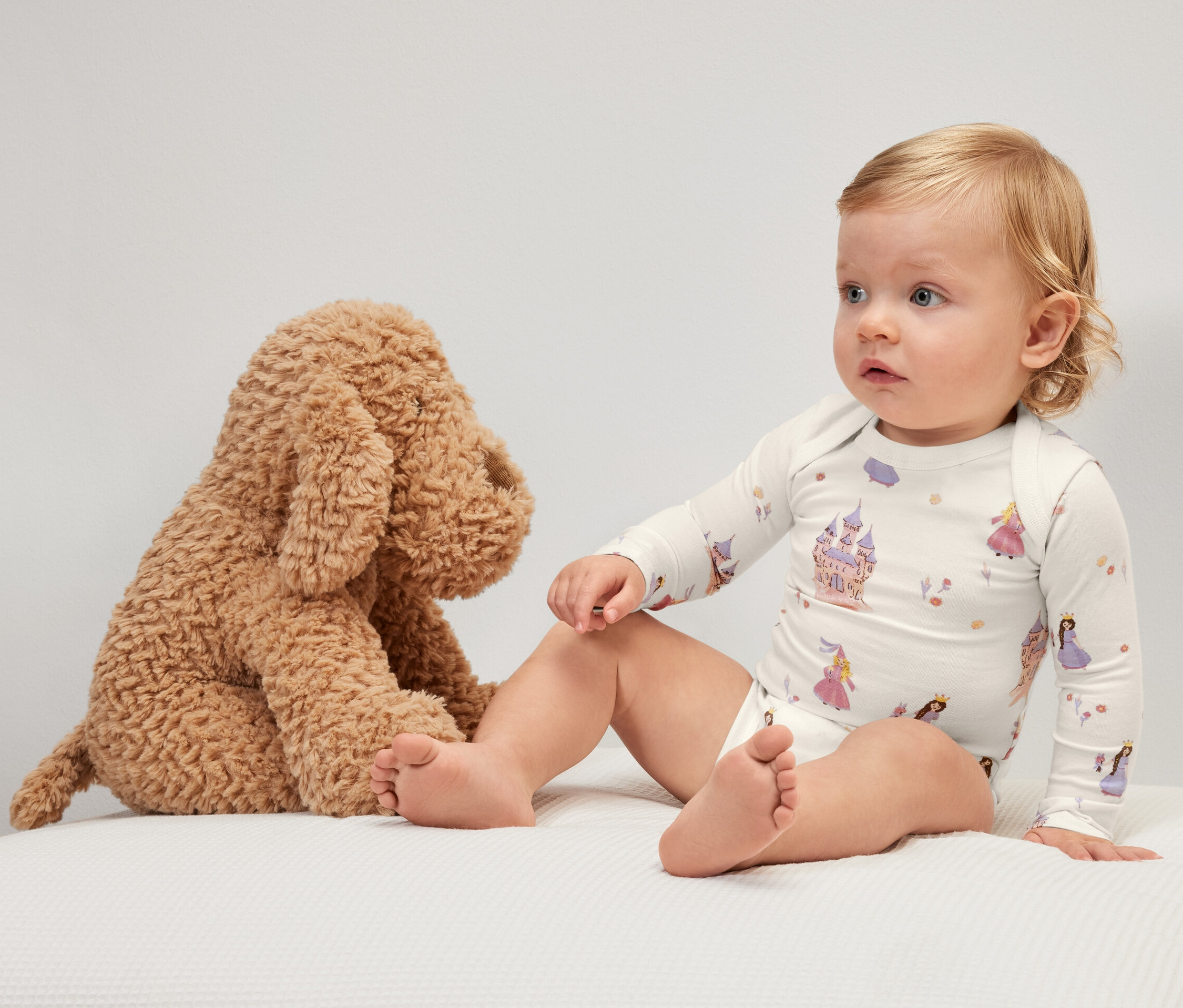 Dziecko siedzi obok pluszowego psa, ubrane w body niemowlęce rosnące z dzieckiem, w księżniczki.