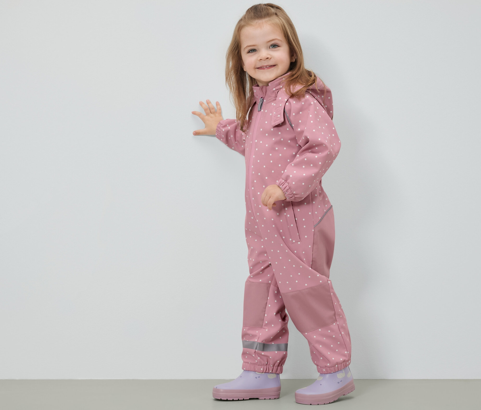 Uśmiechnięta dziewczynka w kombinezonie softshellowym w kropki stoi przy ścianie.