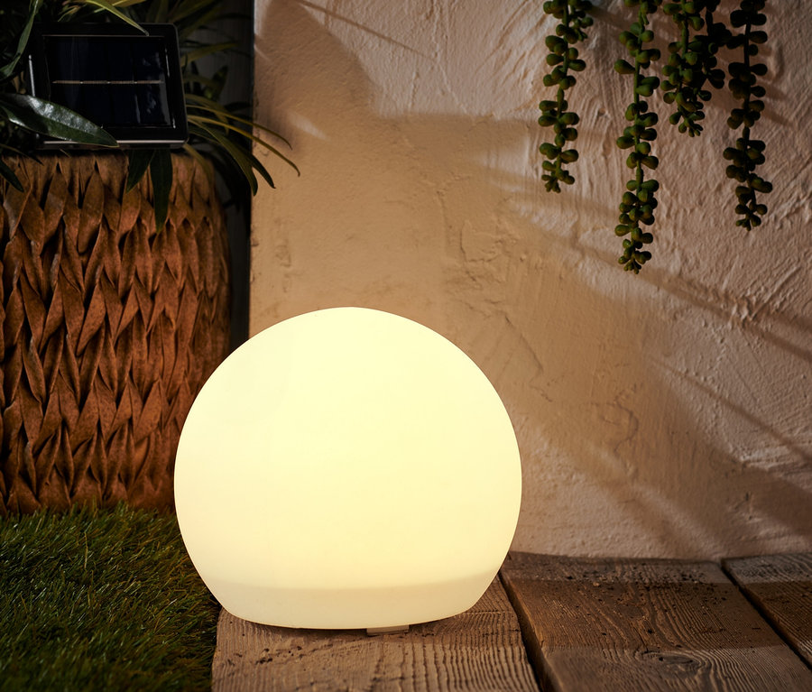 Solarna lampa w kształcie kuli, ok. 20 cm, stoi na drewnianej podłodze obok zielonej rośliny.
