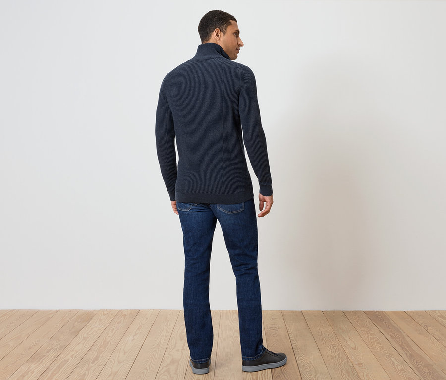 Mężczyzna stoi tyłem do kamery, ubrany w granatowy sweter i dżinsy.