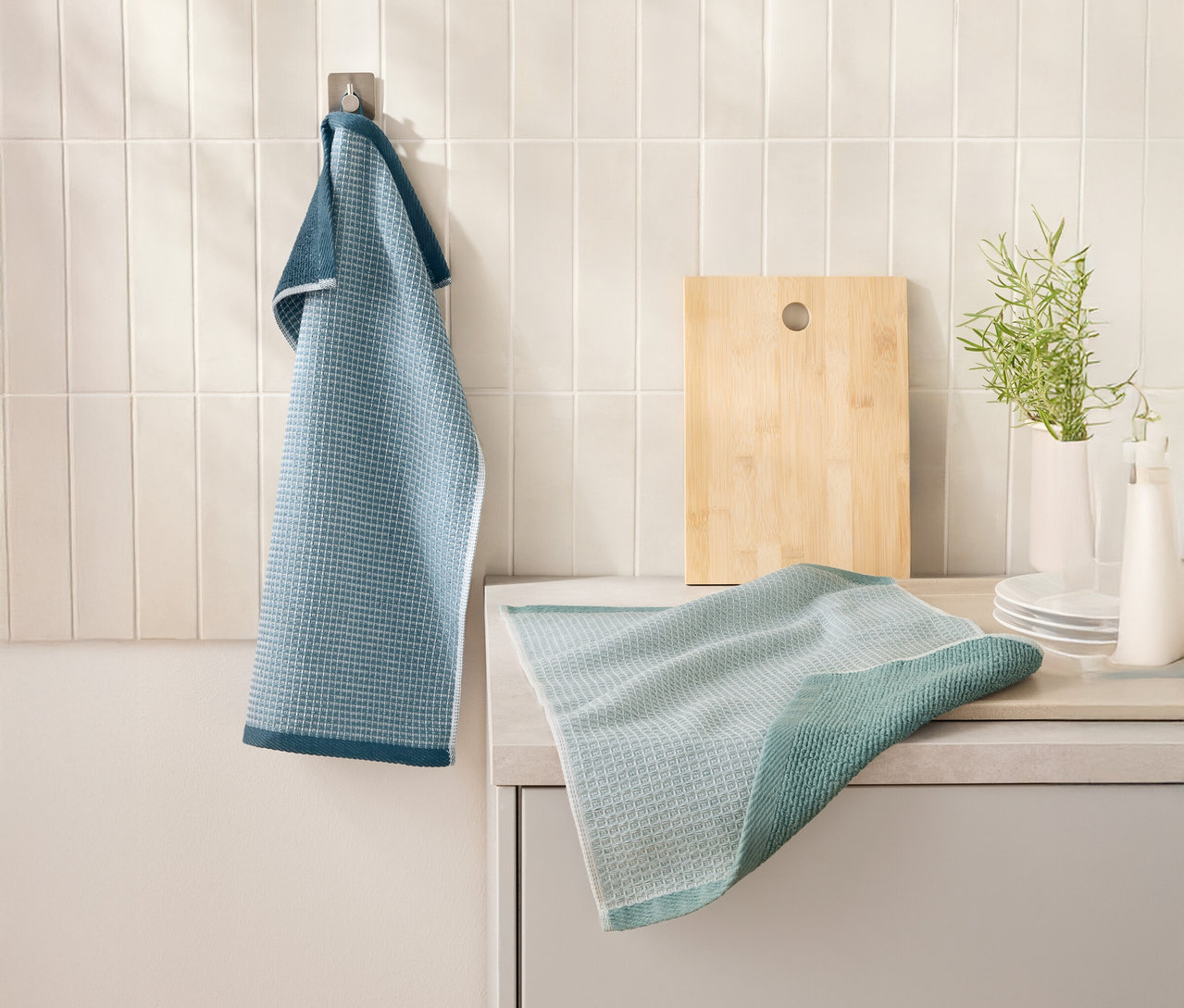 Niebieski 2 Waffelpiqué-Küchenhandtücher wisi na haku obok deski do krojenia. Kolejna ściereczka kuchenna leży na blacie kuchennym.
