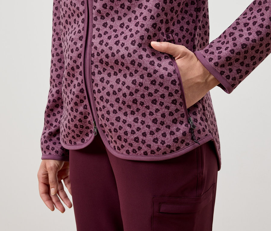 Kobieta trzyma rękę w kieszeni fioletowej kurtki we wzory, noszonej z fioletowymi spodniami.