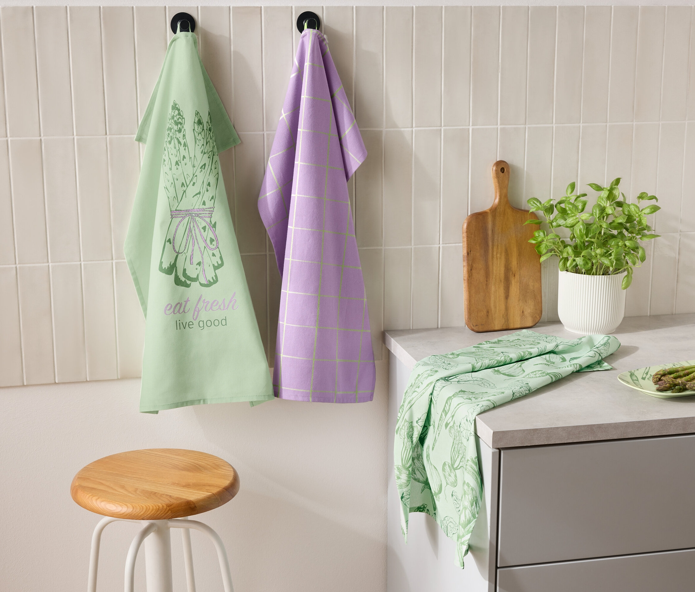 Dwie ściereczki wiszą obok siebie na ścianie, jedna zielona z motywem szparagów i napisem "eat fresh live good", a druga fioletowa w kratę. Na blacie leżą zielona ściereczka i talerz ze szparagami. Obok stoi deska do krojenia i doniczka z ziołami.