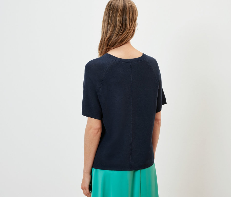 Kobieta tyłem do kamery ma na sobie granatową bluzkę z cienkiej dzianiny i zieloną spódnicę.