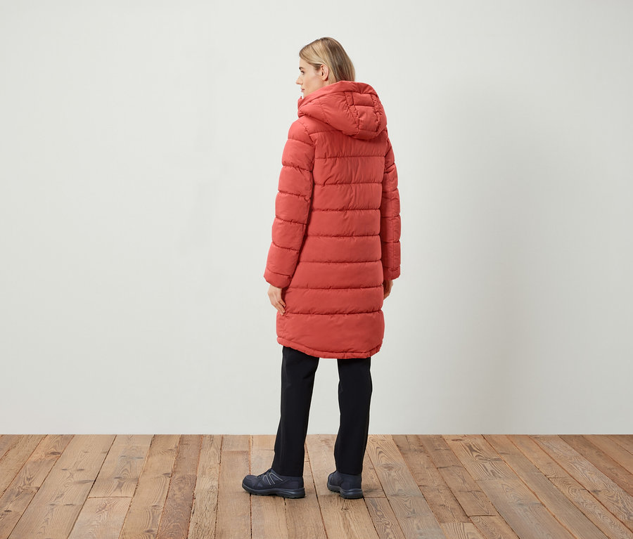 Kobieta stoi tyłem, ubrana w czerwoną, pikowaną kurtkę z kapturem, czarne spodnie i ciemne buty.