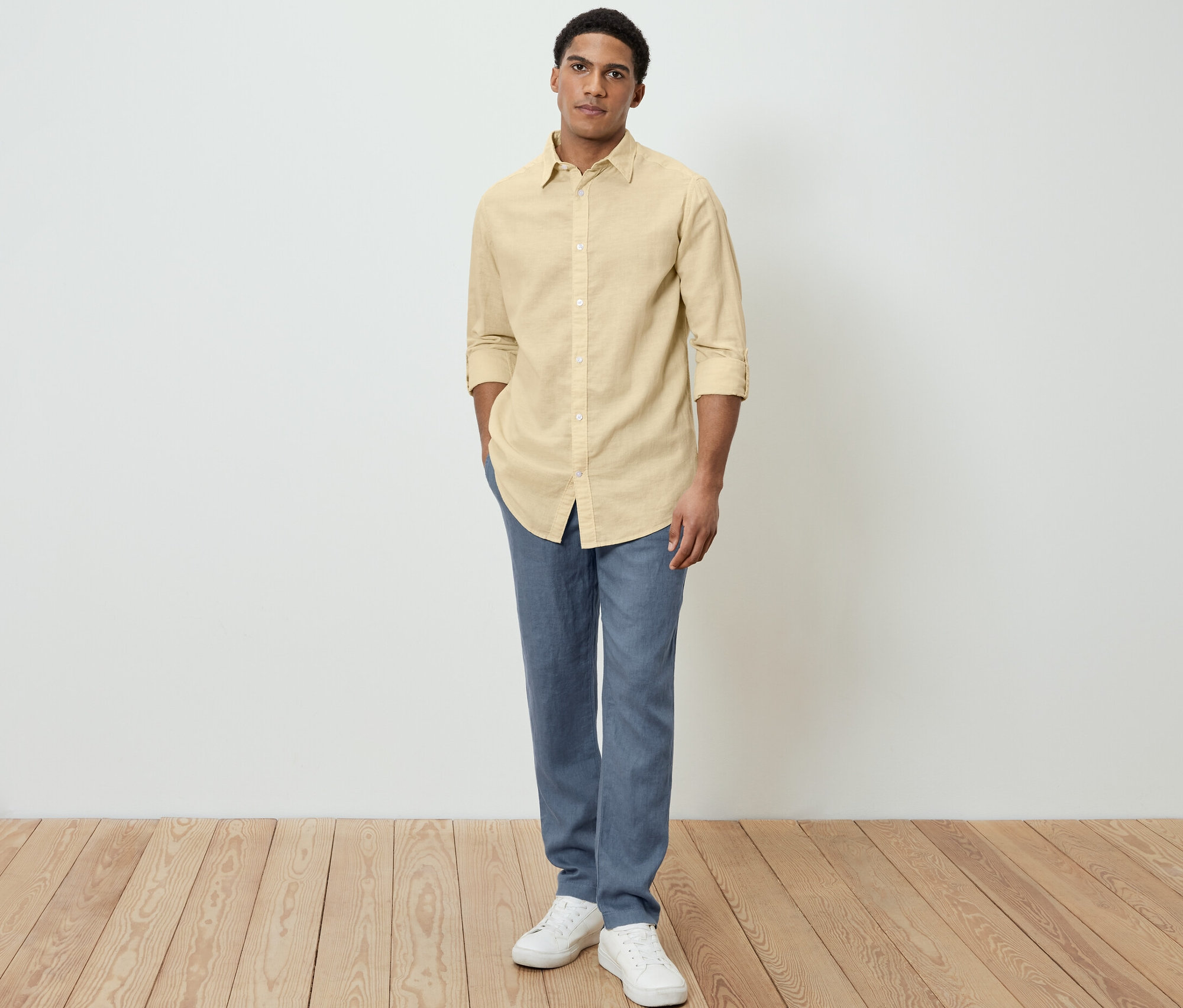 Mężczyzna pozuje w żółtej koszuli z lnem, niebieskich spodniach i białych butach.