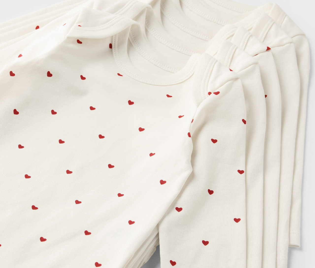 Stos pięciu białych body rosnących z dzieckiem, z czerwonym wzorem w serca.