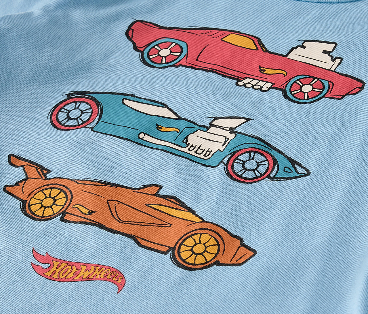 Na jasnoniebieskiej koszulce znajdują się trzy narysowane samochody Hot Wheels w kolorach czerwonym, niebieskim i pomarańczowym.