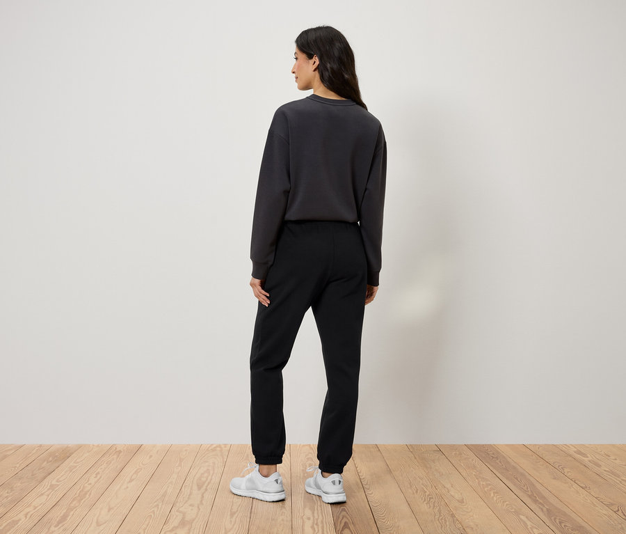 Widok z tyłu kobiety w czarnych spodniach dresowych, szarej bluzie i białych butach.